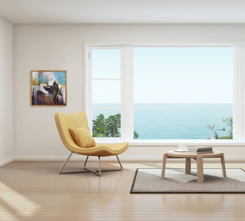 Insitu image of All Aboard, a mixed media abstract painting by Kathryn Gruber. The room has a large window looking out onto the blue ocean, and inside the room, a yellow chair and a coffee table on a rug. The floor is timber, and the painting is hung on the white wall, to the left of the large window.