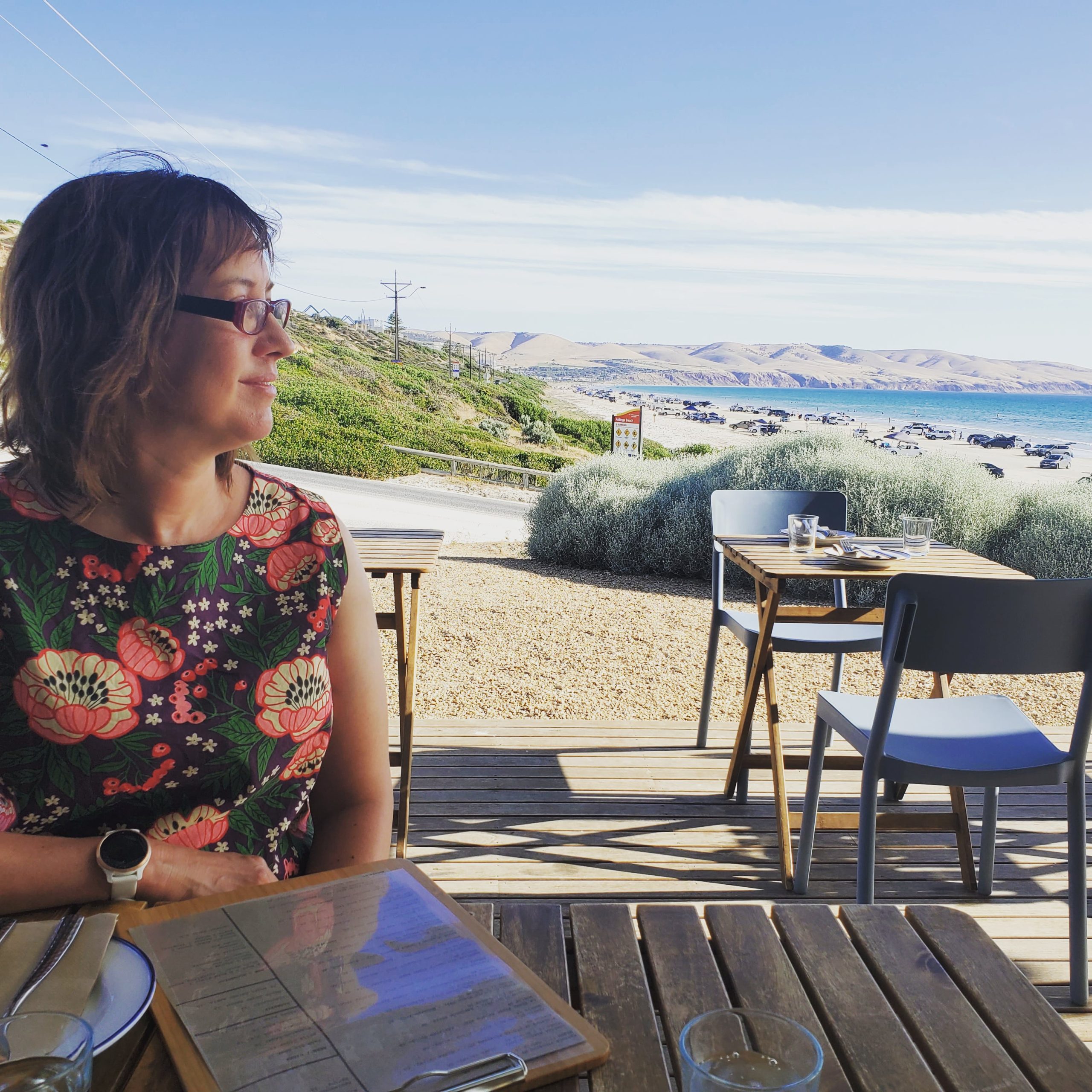 Woman with colourful top looks out over the coast whilst sitting at a cafe table.