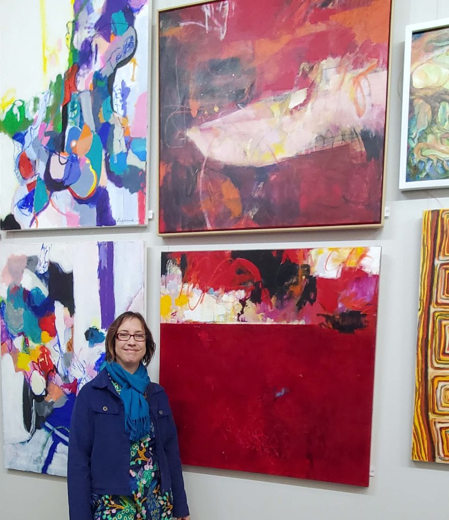 Artist Kathryn Gruber stands, smiling in front of two of her large abstract paintings at the RSASA Abstract Art Show finalists exhibition